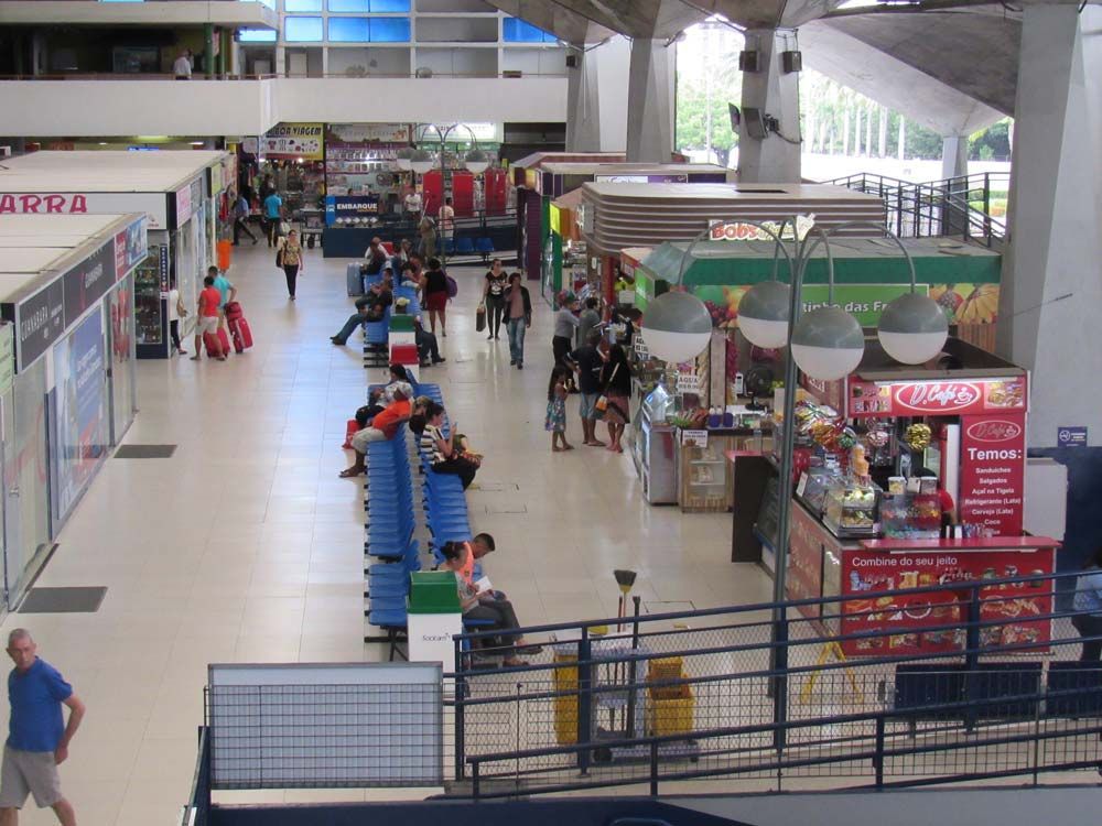 Interior de aeroporto com pessoas caminhando em frente a lojas e quiosques, estrutura de concreto, iluminação forte.