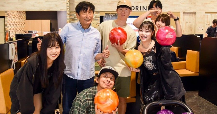 Group of people at a bowling alley holding bowling balls. They smile and pose for the camera, illuminated by bright lights.