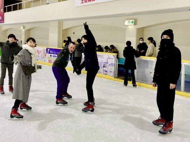 People ice skating in an indoor rink. Some are bundled up, one raises their arms, others wear masks.