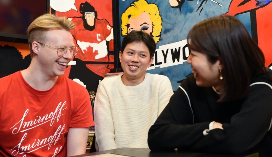 Three people laughing in front of a colorful wall; one wears a red shirt with white text, another a white sweater, and the third a black jacket.