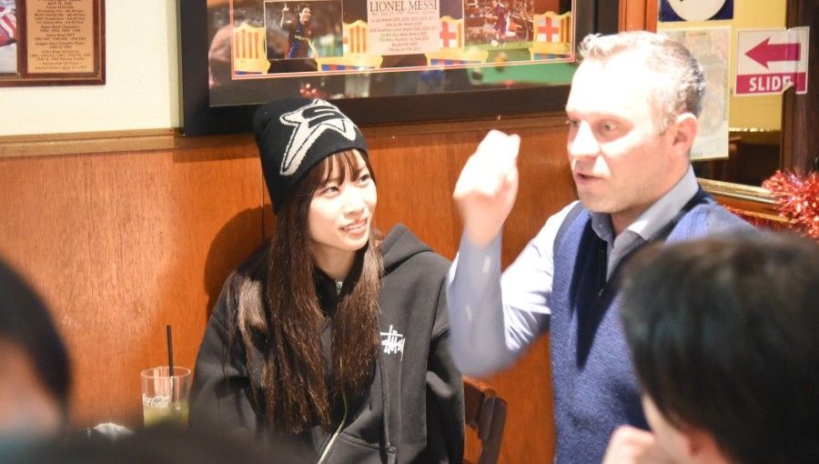Woman in beanie and man in vest conversing at a table. Man gesturing with his hand. Interior setting, restaurant.