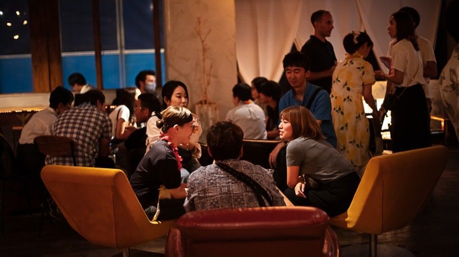 People gathered in a warmly lit lounge; several are conversing, some in traditional clothing.