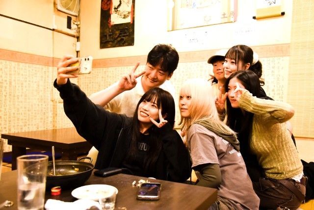 Group of people taking a selfie in a dimly lit restaurant, smiling and making peace signs.