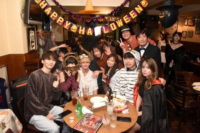 Group of costumed people at a Halloween party, posing around a table with drinks and snacks, banner says 
