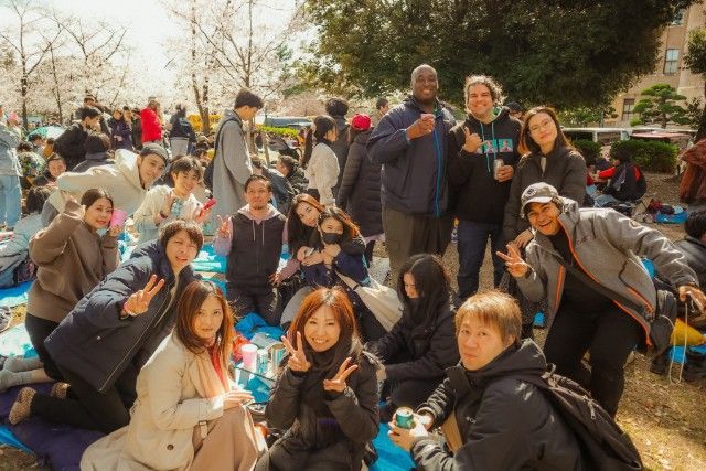 Group of people picnicking outdoors under cherry blossom trees, making peace signs and smiling.