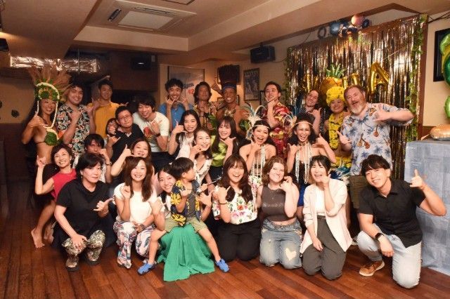Group of people at a Hawaiian-themed party, smiling and posing for a photo indoors with decorations.