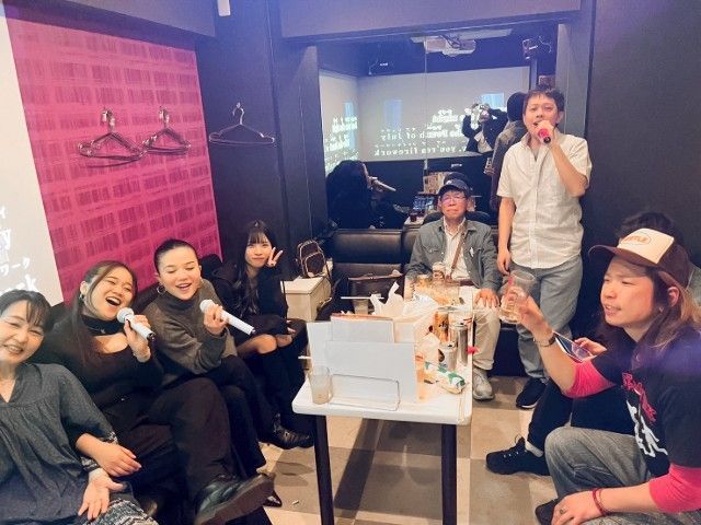 People singing karaoke in a room; a few are holding microphones. White table with drinks.