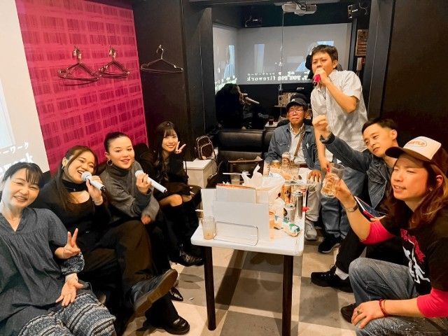 Group of people singing karaoke in a dimly lit room, some holding microphones, with a table of drinks in front.