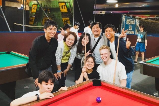Group of people smiling at a pool table, inside a lit recreational space.