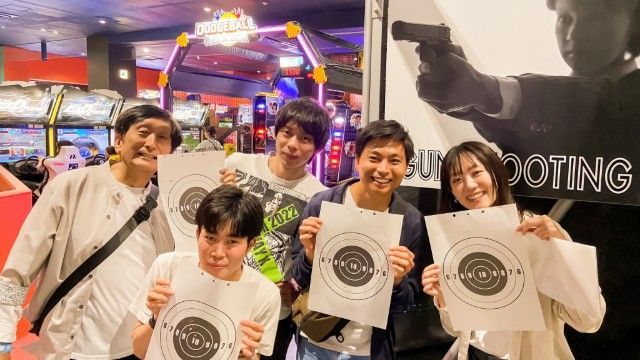 Five people holding target sheets in an arcade, smiling. Behind them are game machines.