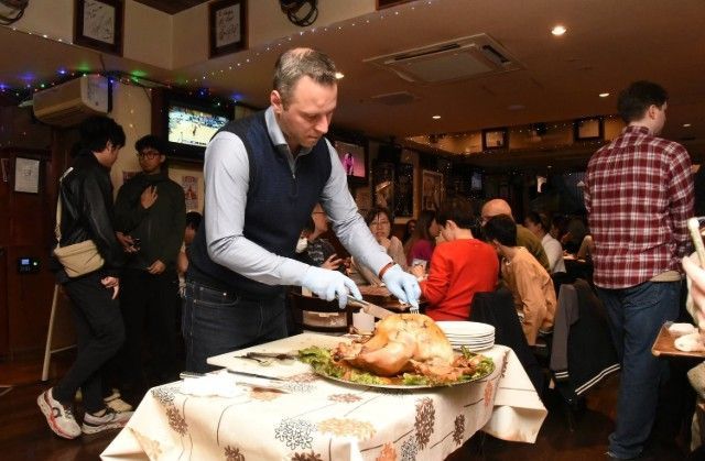 Man carving a roasted turkey at a table, inside a crowded restaurant. Other people observe.