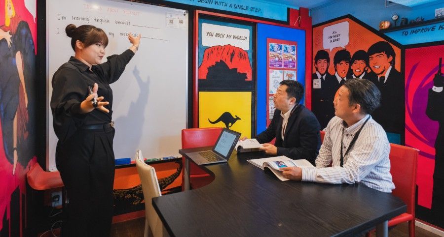 A person instructs two seated individuals at a table in a brightly colored room decorated with posters.