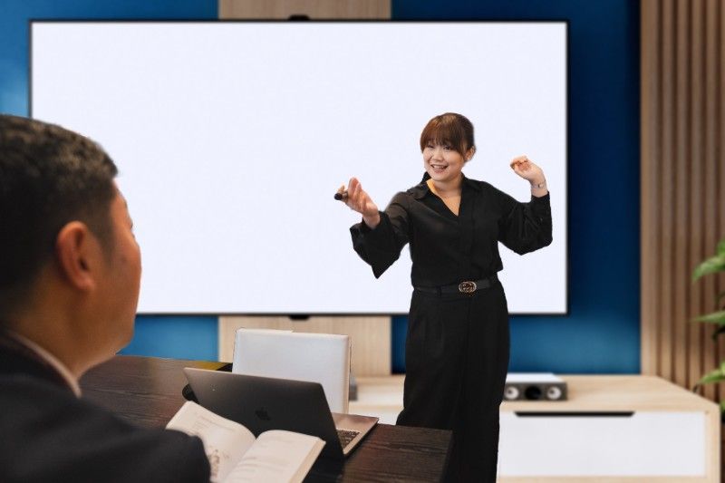 ビジネスミーティングでジェスチャーを交えながらプレゼンテーションをする女性。背後には何もないスクリーンがあり、テーブルにはノートパソコンを持った人物が座っている。