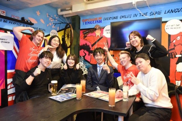 色とりどりの看板や飲み物がテーブルに並べられた、活気あふれるゲームセンターやゲーム発売イベントで、笑顔の人々がポーズをとっている。
