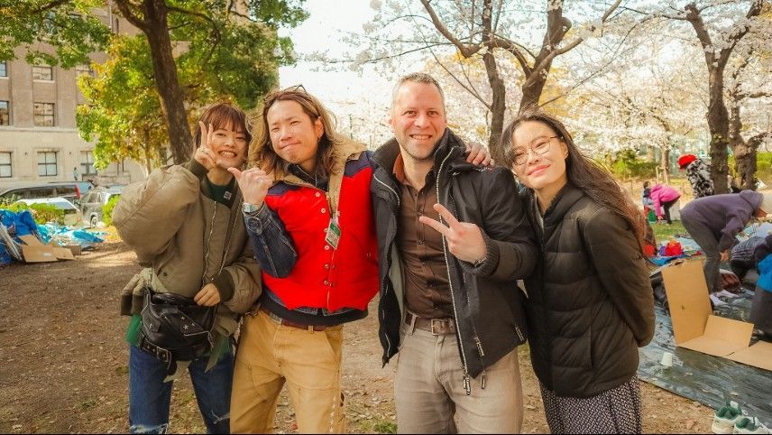 Four people smiling, posing outdoors. Cherry blossoms and buildings in the background. Two give peace signs.