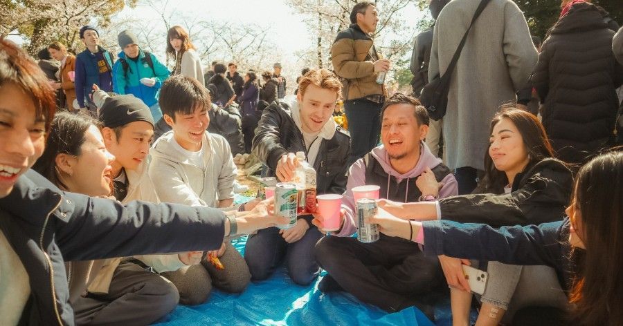 People toasting with drinks while seated on a blue tarp in a park, blurred background of others.