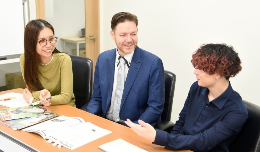 Three people sitting around a table in an office setting, engaged in a professional discussion with papers spread out.