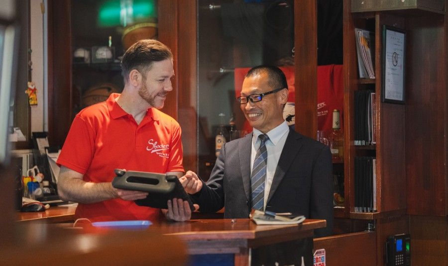 A person in a red shirt shows a tablet to a professional in a suit across a wooden counter in an office setting.