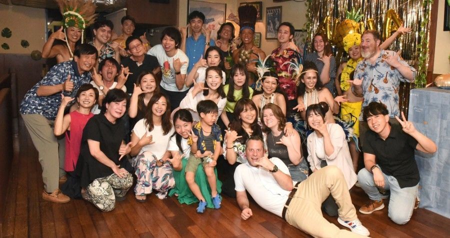 Group of people smiling, posing for a photo indoors. Some making peace signs, celebratory expressions. Wooden floor, festive backdrop.