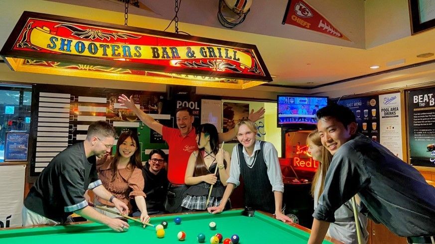 People at a pool table in a bar called Shooters. Smiling, raising hands, playing pool. Illuminated sign, TV.