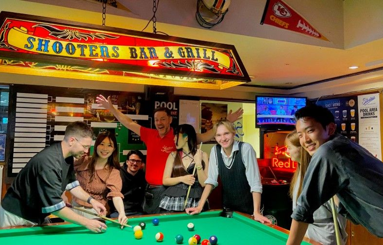 ライトアップされた「シューターズ・バー＆グリル」の看板の下、バーの中で笑いながらビリヤードを楽しむ友人グループ。