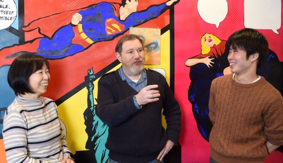Three people smiling and talking in front of a colorful comic book mural featuring Superman.