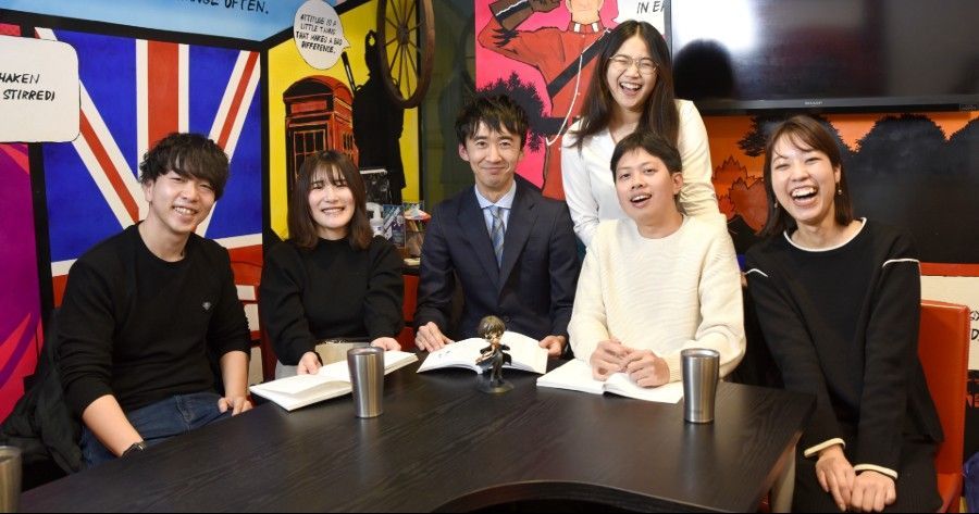 Six people sit around a table, laughing. The setting includes a Union Jack flag and colorful decor.