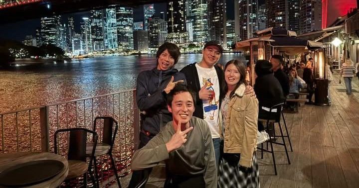 Four friends posing by a riverside at night with city lights reflecting on the water.