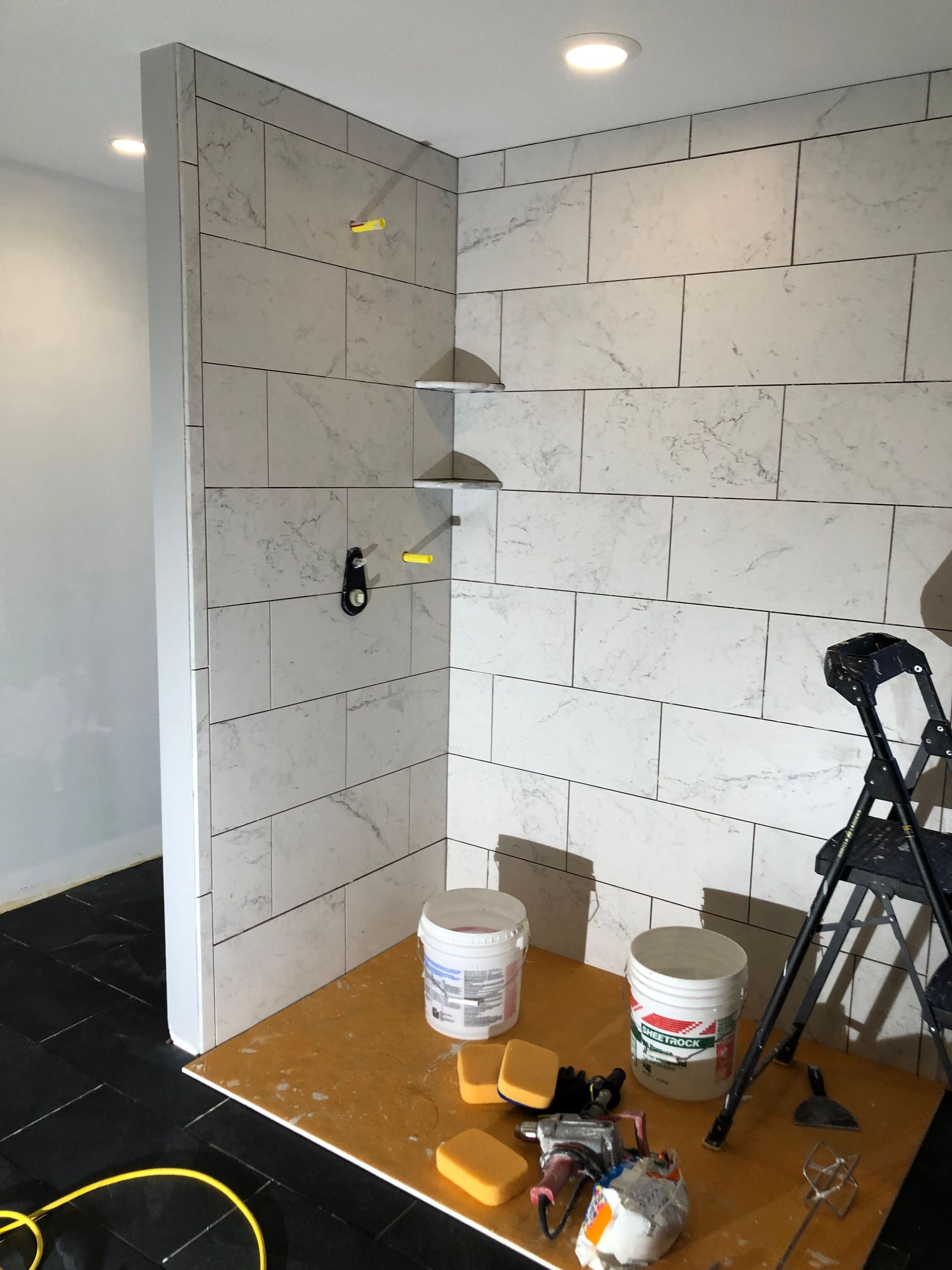 Bathroom corner with tiled walls, shelves, buckets, tools, and a stepladder.
