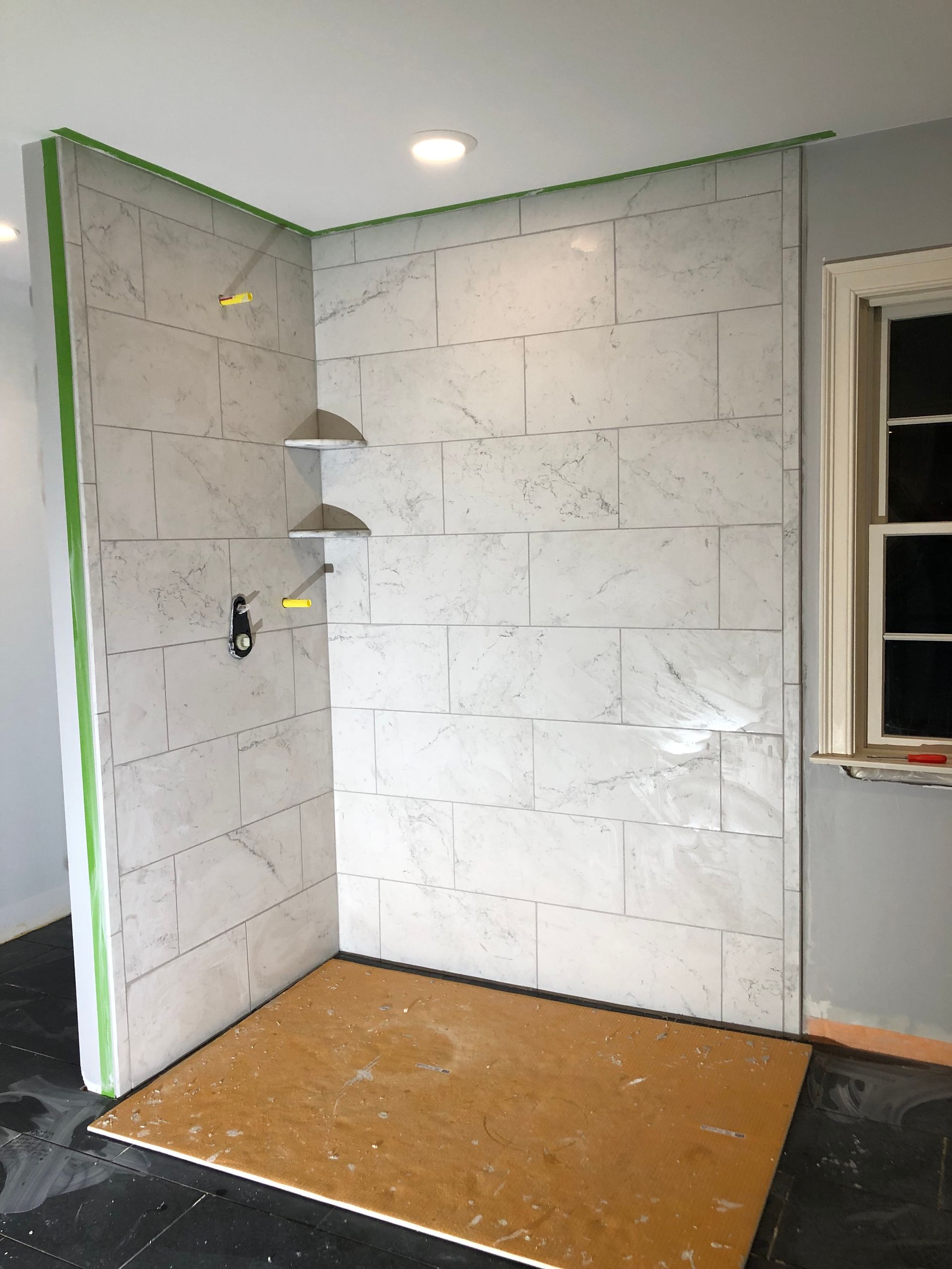 Shower walls tiled with marble-like stone, two built-in shelves, wood floor, window on right.