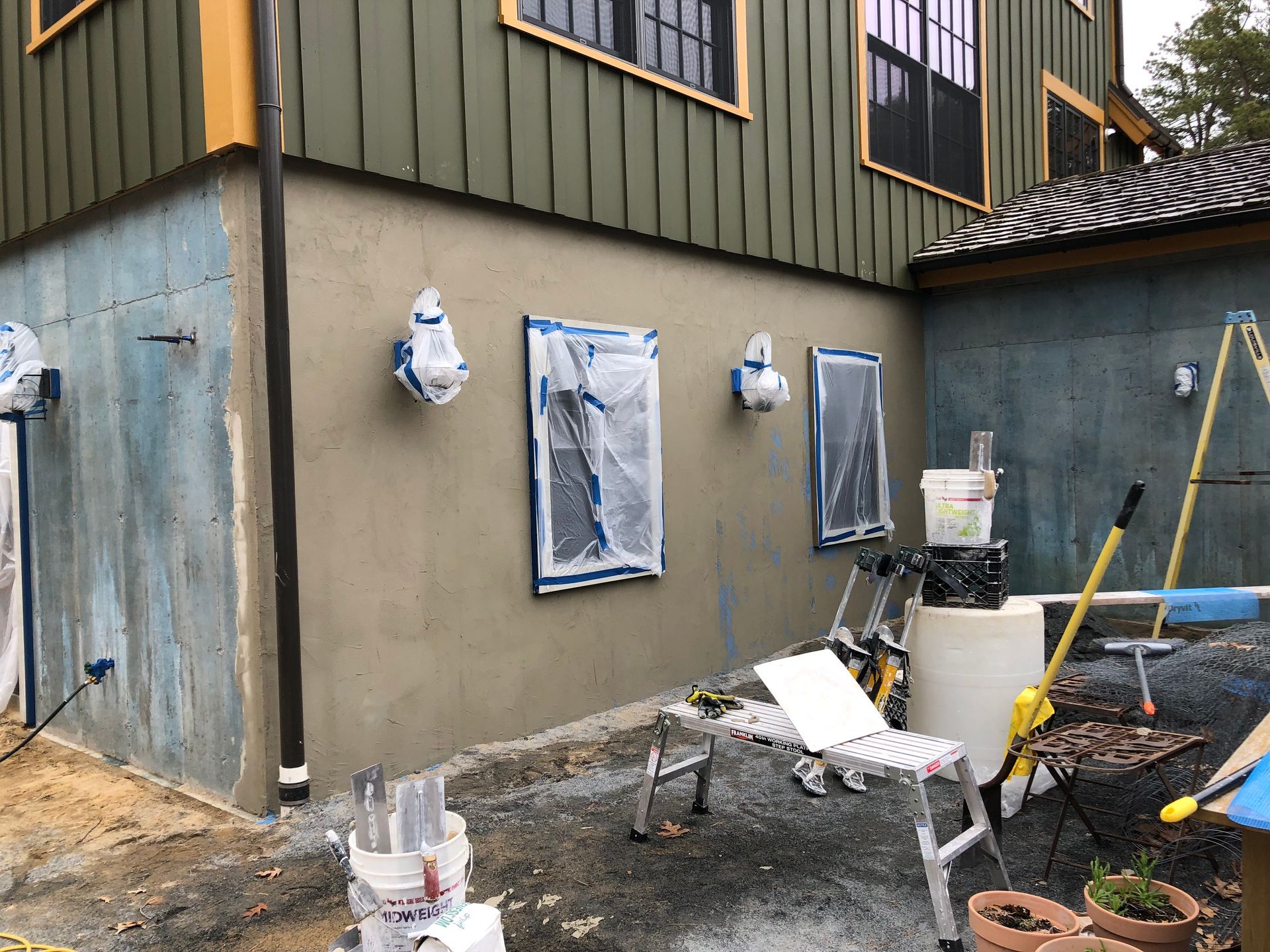 Exterior house wall under renovation; windows taped. Blue and tan stucco. Ladder present.