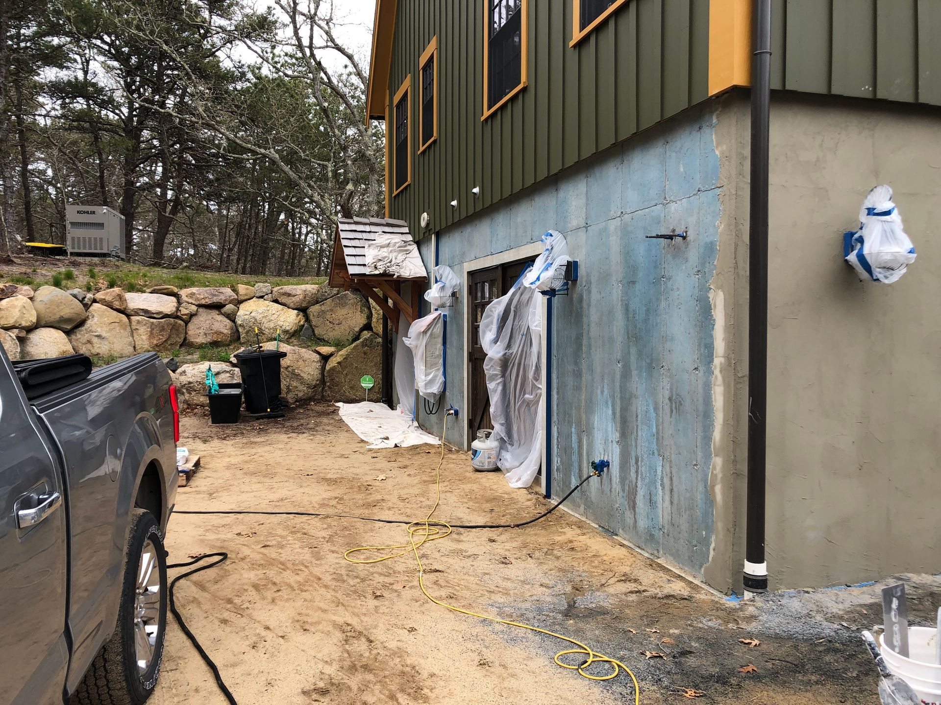 A house exterior under construction; light blue wall, covered door, gray truck, wooden trim and landscaping.