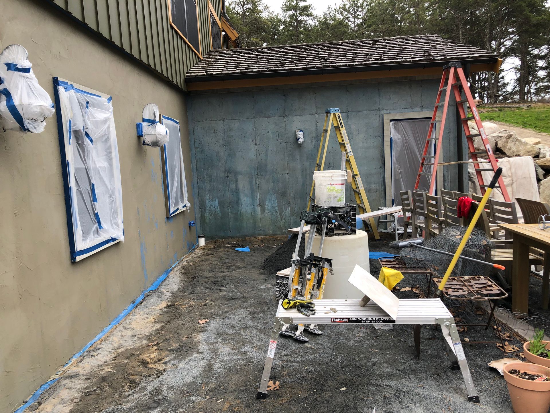 Exterior wall construction site: taped windows, blue stucco, ladder, tools, gravel ground, and wooden structure.