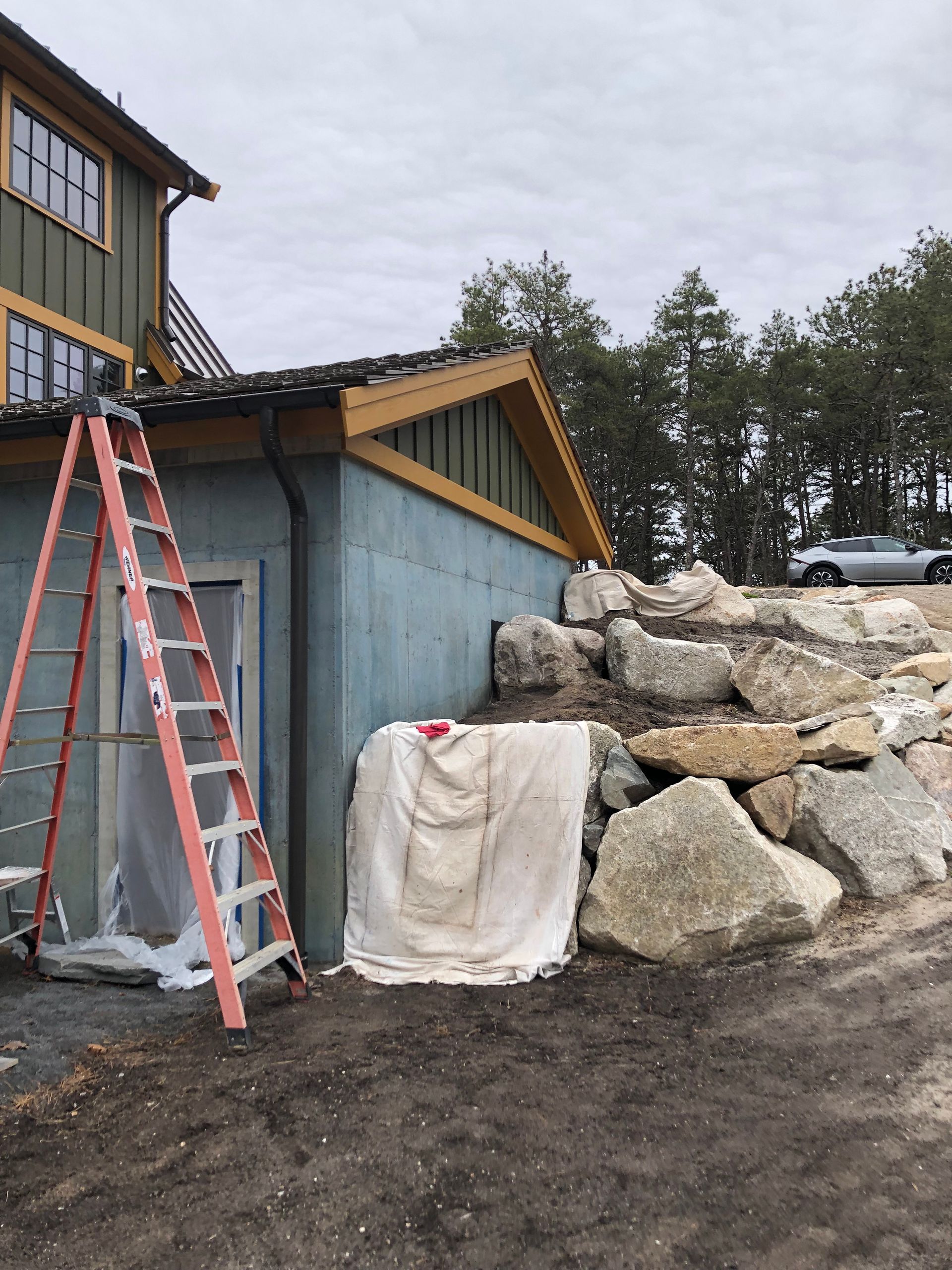 House exterior with ladder, tarp-covered wall, and rock retaining wall; overcast sky.