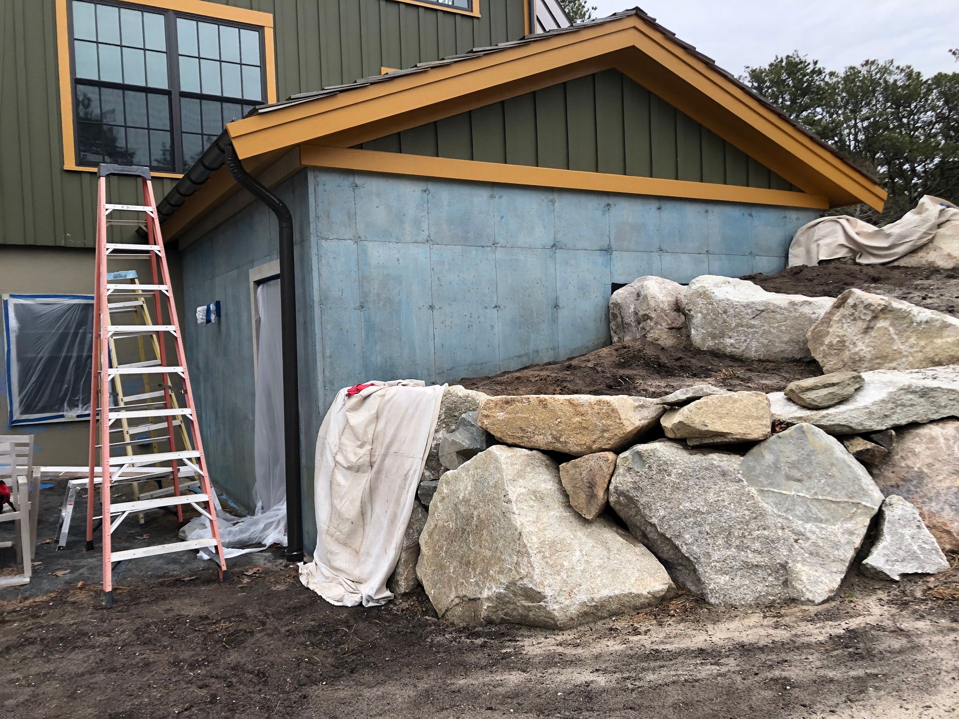 A building under construction with a rock wall, ladder, and a door. Blue insulated walls and green siding.