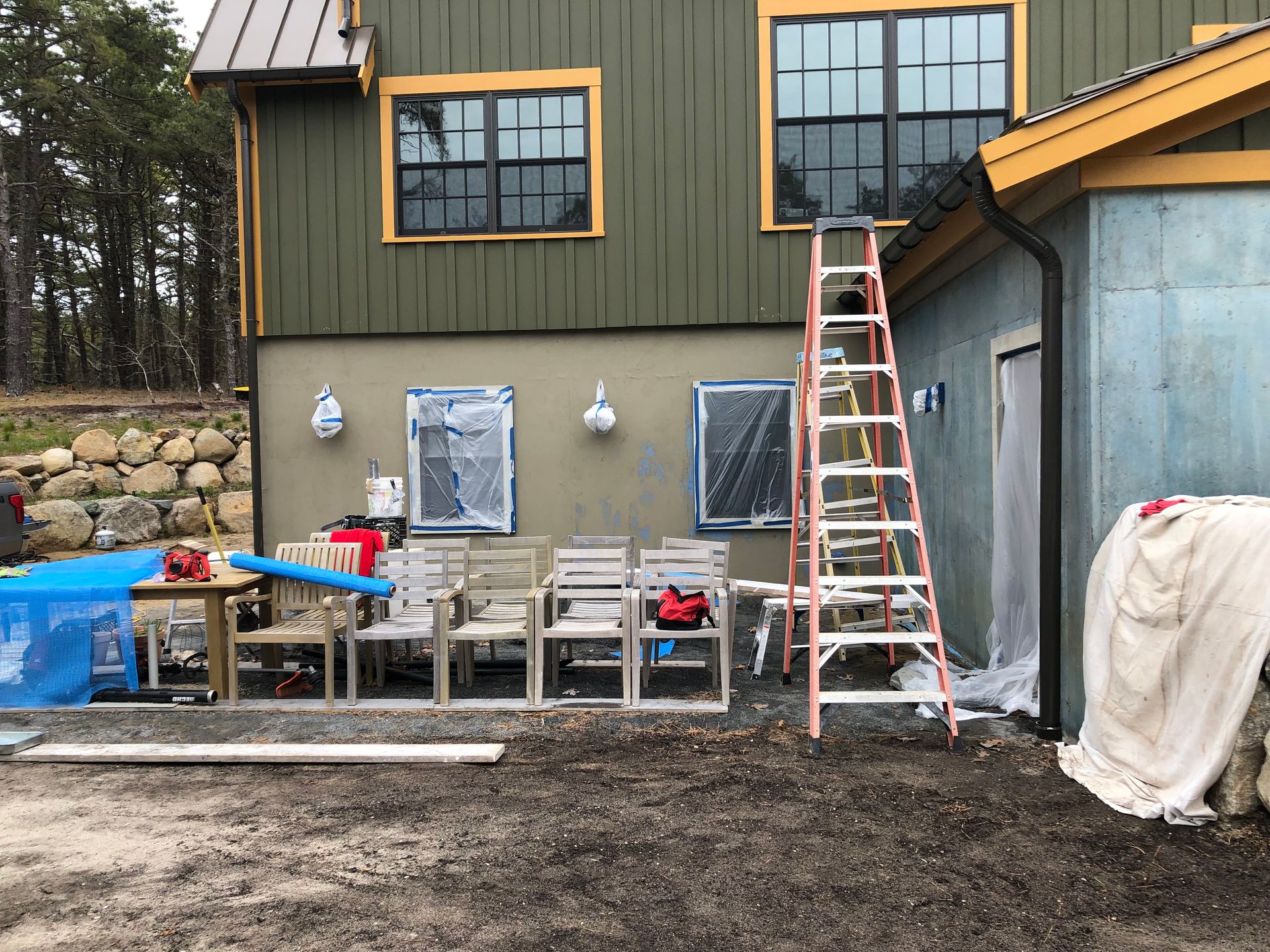 Exterior of a house under construction; ladder against wall, windows covered, chairs, tools, and materials on the ground.