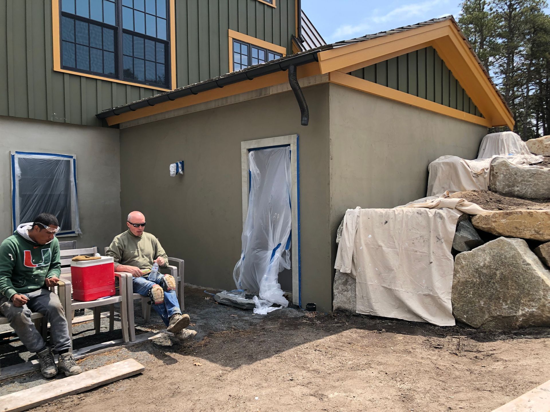 Two men sitting outside a building under construction. Grey plaster walls, tarp-covered door, rocks, and a green house.