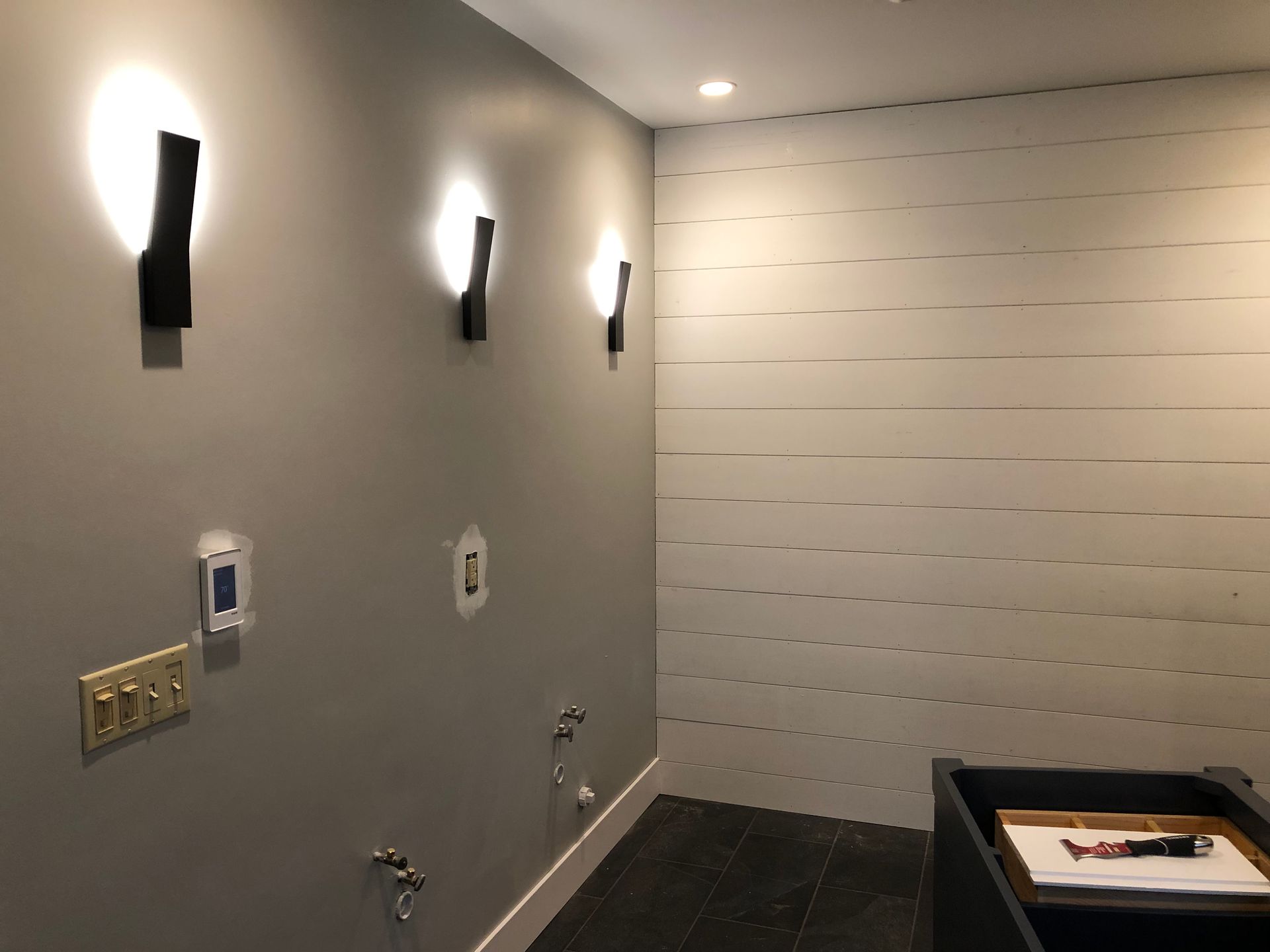 A gray room with three black sconce lights on the left wall and a white, horizontal plank wall on the right.