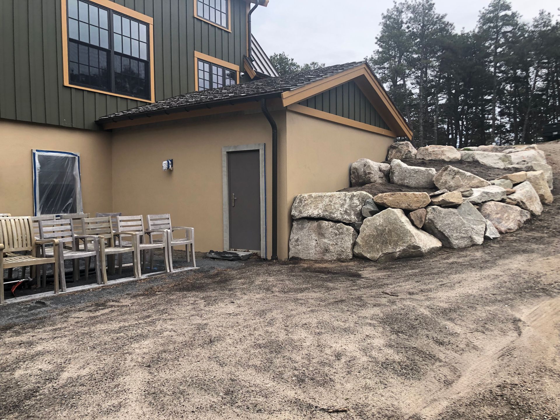 Exterior of a building with a rock retaining wall and gravel ground.  Chairs sit on a patio area.