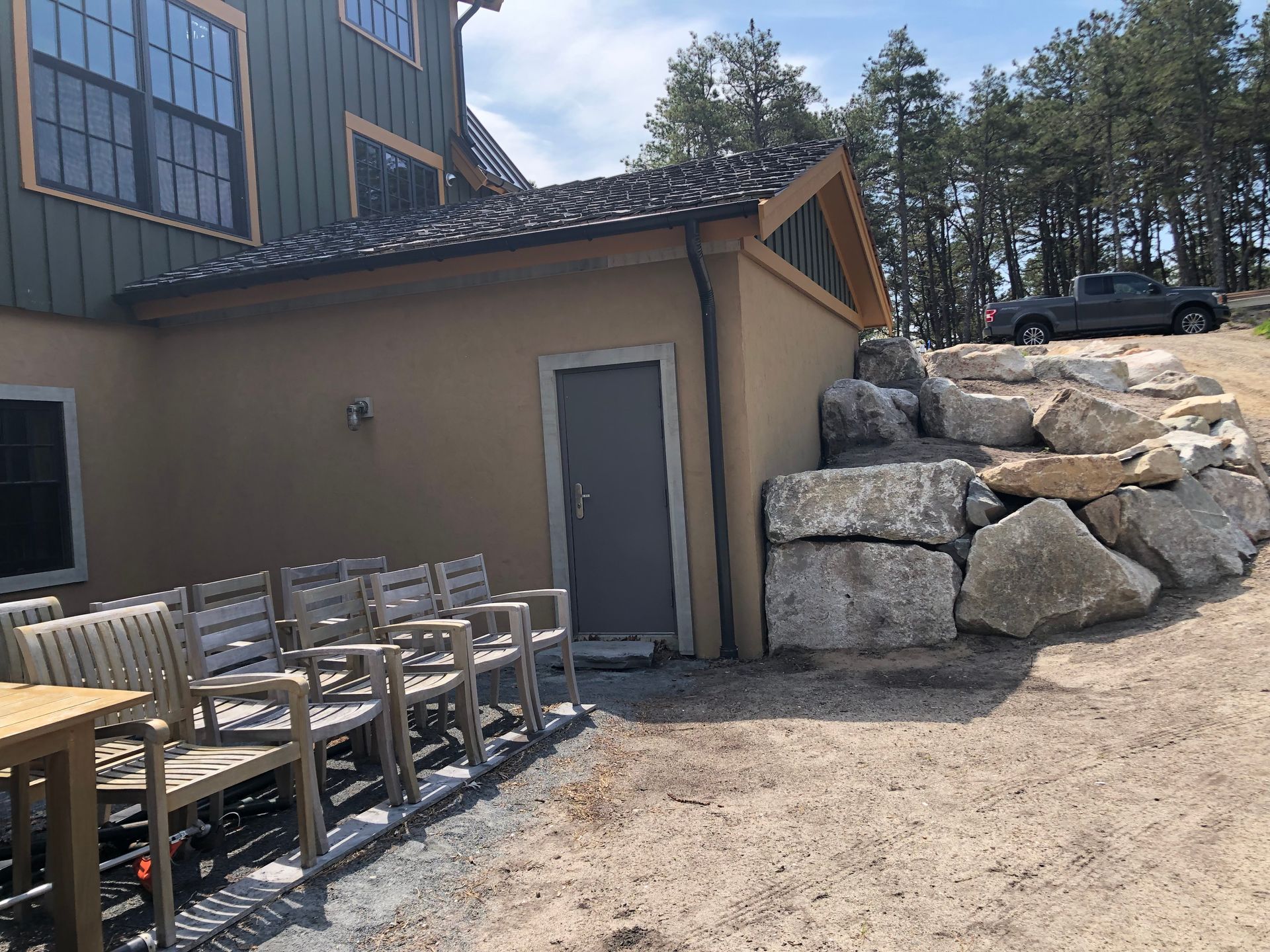 Building with a tan wall, gray door, and wooden chairs outside. Stone steps lead up to a gray truck.