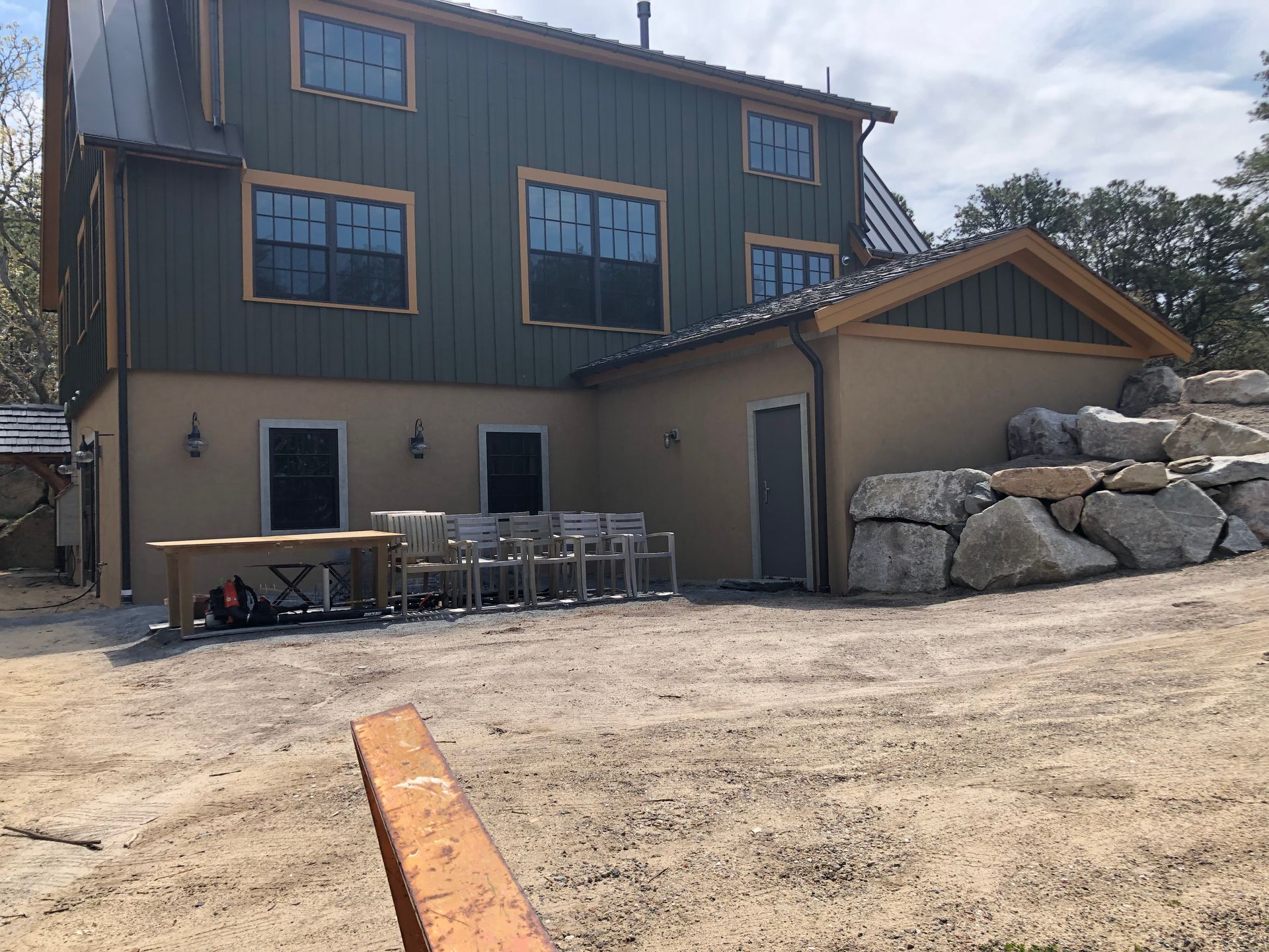 Back of a two-story building with green siding and tan stucco. Outdoor seating area on gravel, with boulders.