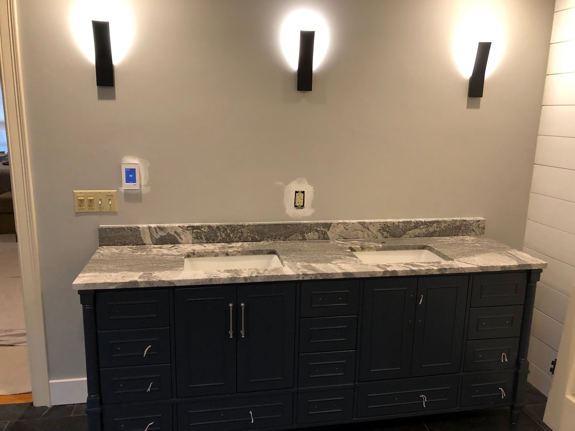 Bathroom vanity with dark blue cabinets, granite countertop, two sinks, three black sconces on a gray wall.