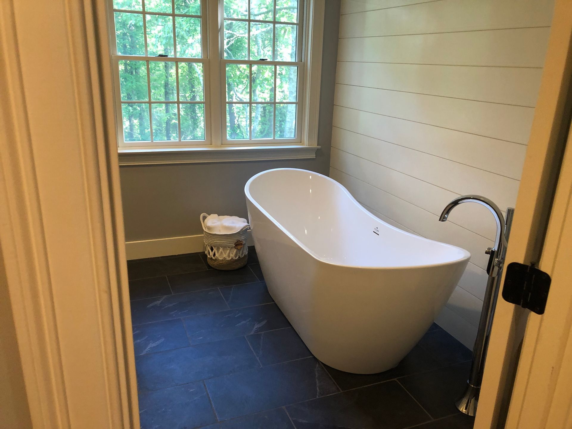 Freestanding white bathtub in a bathroom, with a window and dark tile floor.