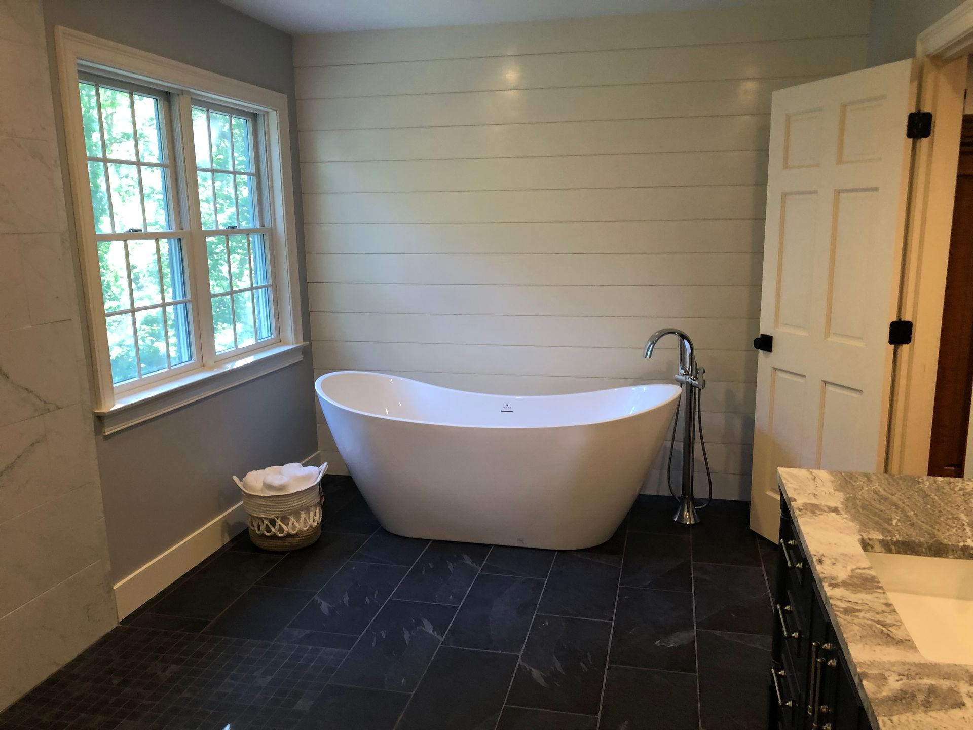 Modern bathroom with a white freestanding tub, wood-paneled wall, dark tile floor, and a window.