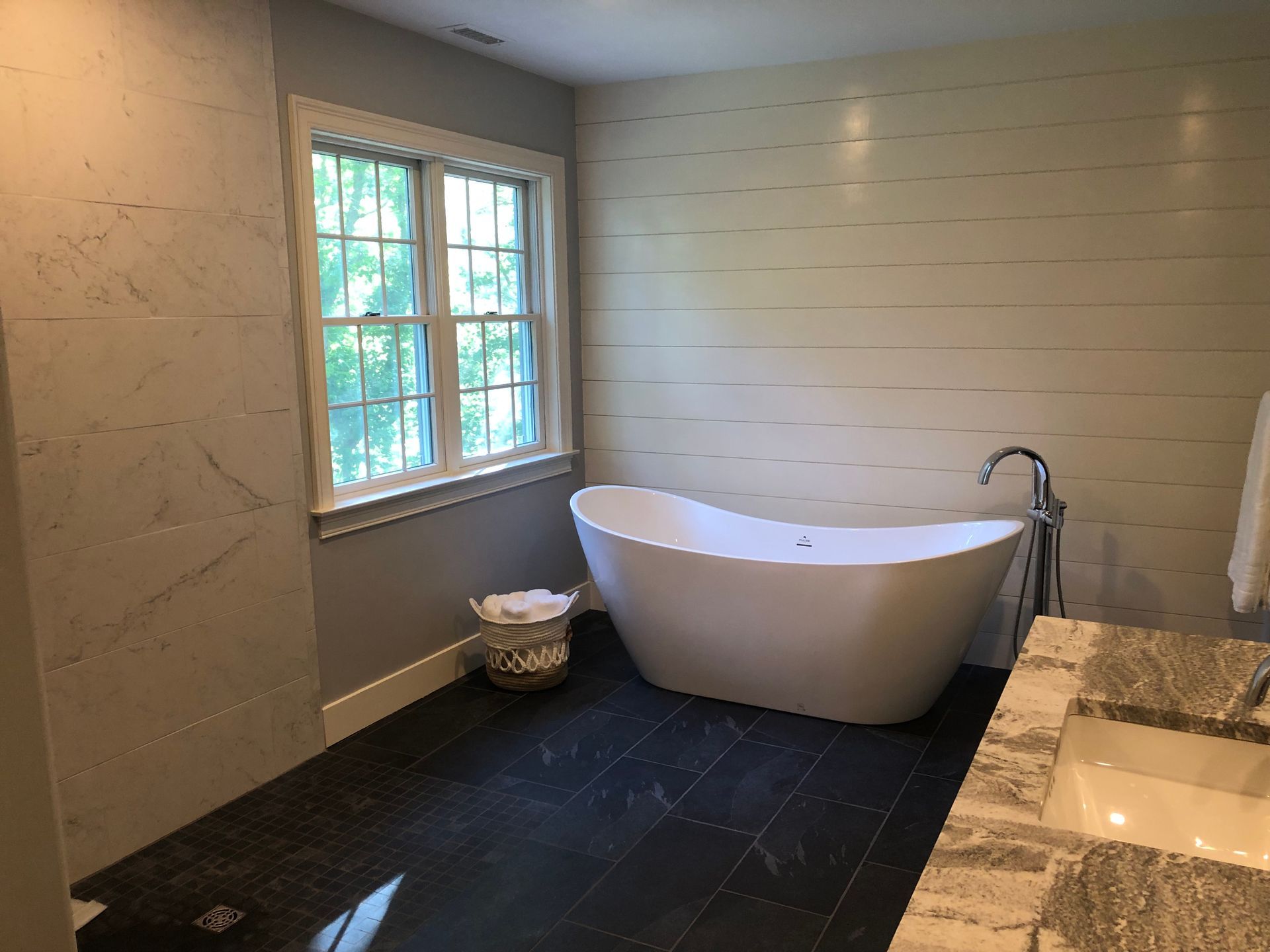 Bathroom with a white freestanding tub, window, dark tiled floor, and marble walls.