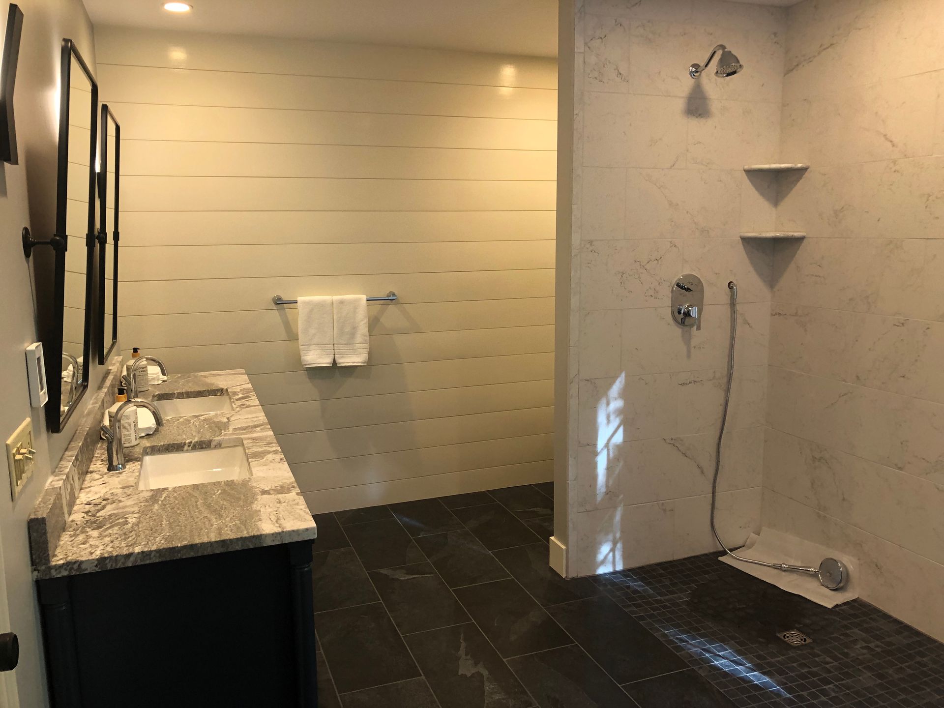 Bathroom with dual sinks, shower, dark tile floor, light wall paneling, and a towel rack.