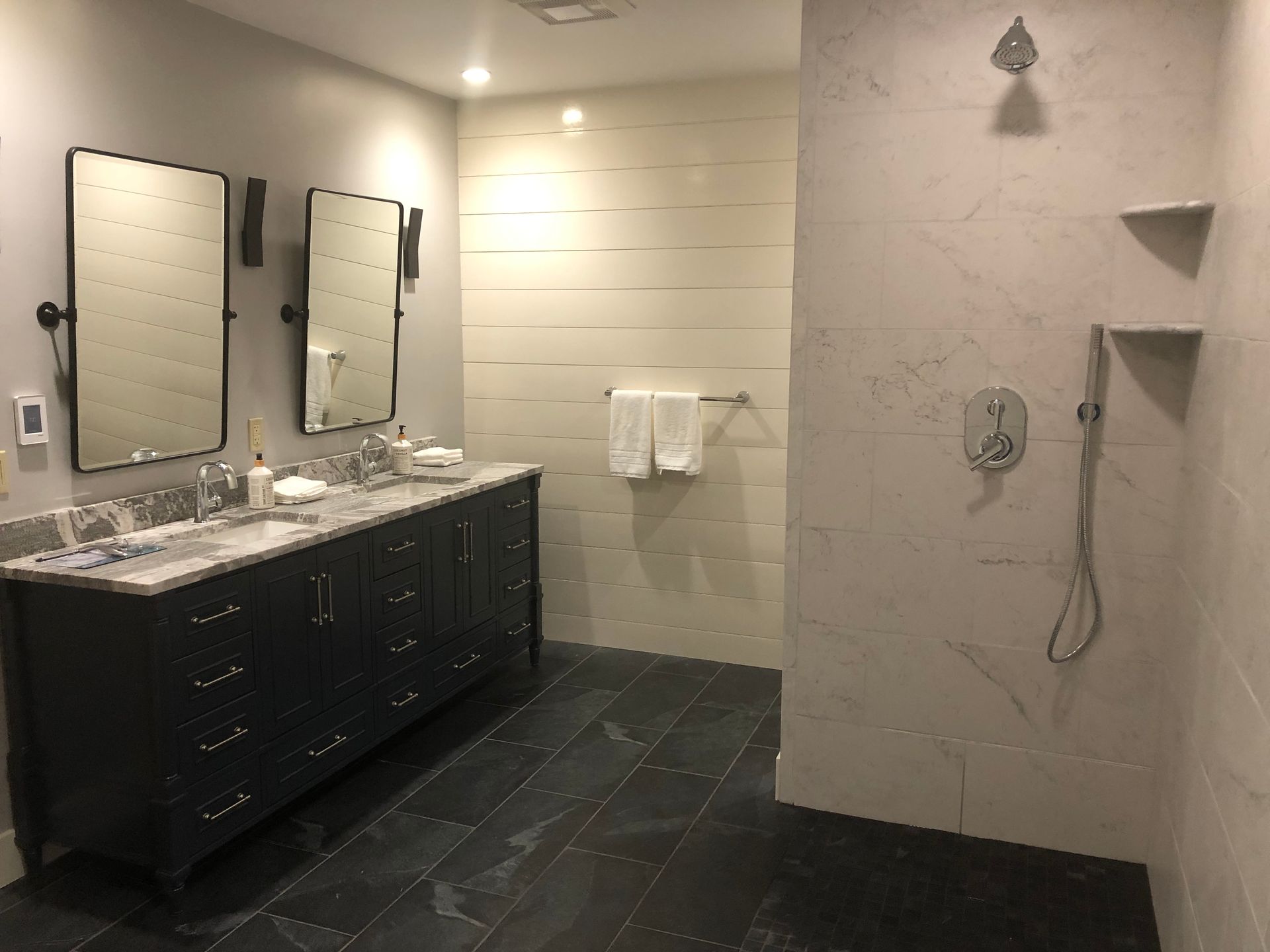 Bathroom with dark vanity, marble shower, and two rectangular mirrors. White wall with wood panels. Dark tiled floor.