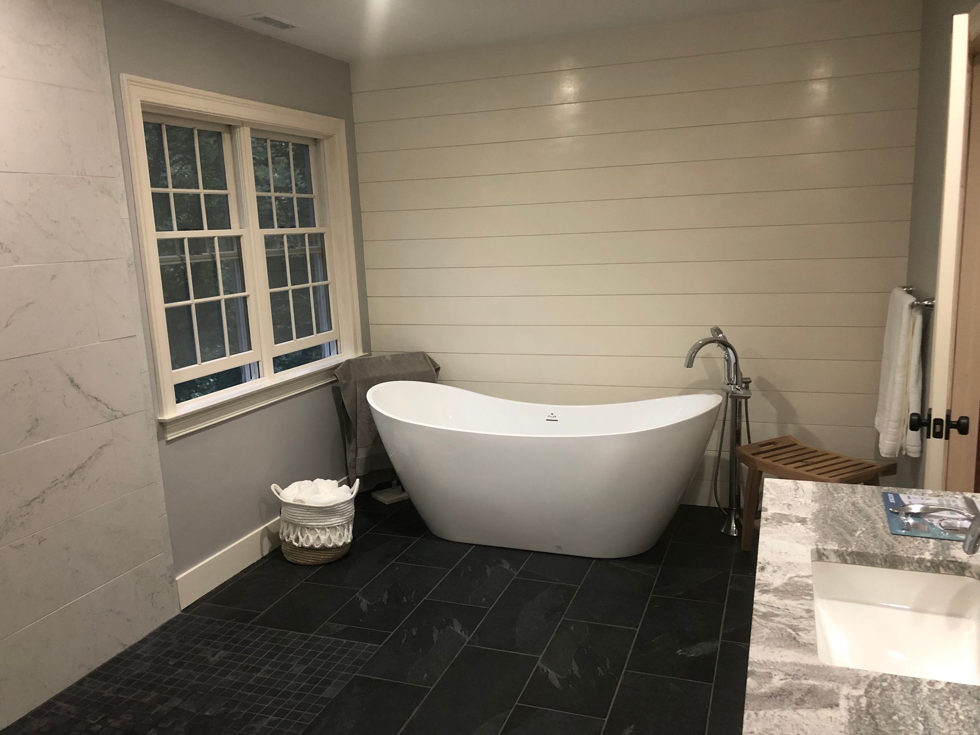 A modern bathroom with a white tub against a shiplap wall, dark tile floor, and a window.