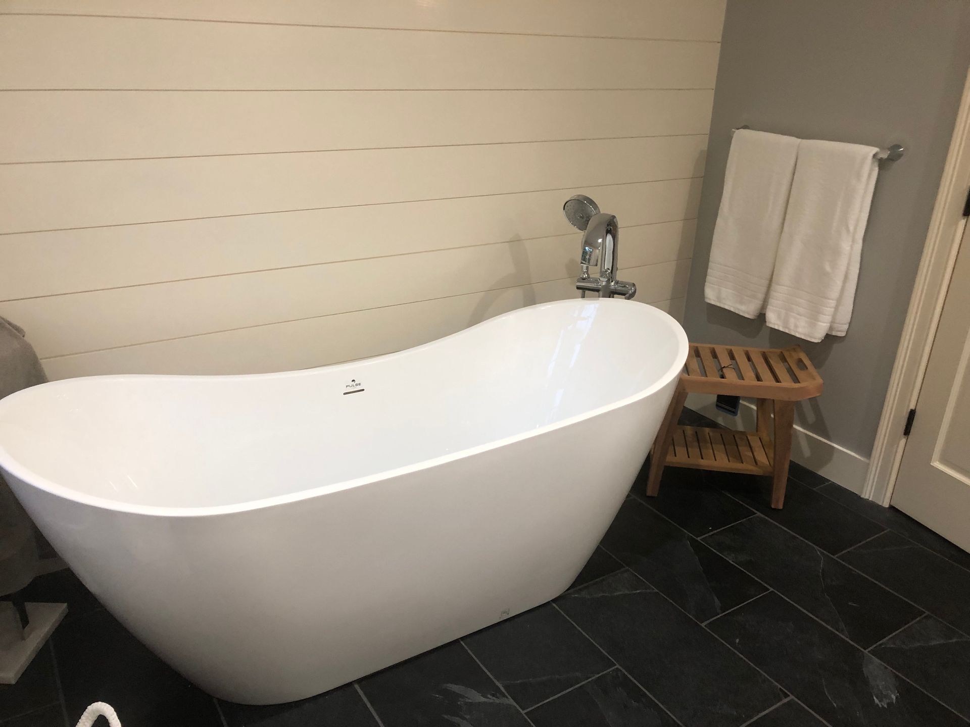 Freestanding white bathtub in bathroom, with hand shower, wooden stool, and white towels.