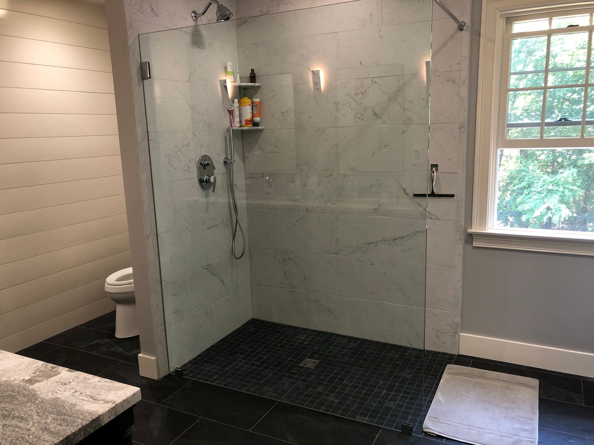 Modern bathroom with glass shower, marble tile, and black pebble floor, next to a toilet and window.